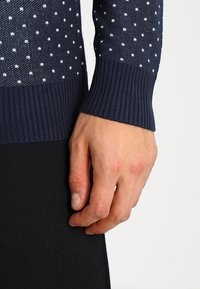 Marinblå tröja med vita prickar, ribbade ärmslut och strukturstickad. Hand som vilar på en svart texturerad tygbakgrund.