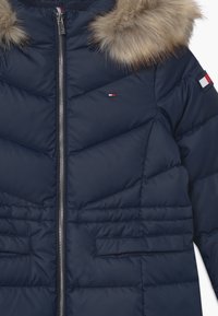 Námořnická péřová bunda s kapucí lemovanou kožešinou, zapínáním na zip, vzorem včelího úlu a logem na rukávu. Měkká textura.