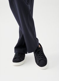 Black suede sneakers with white rubber soles and black laces, worn with loose-fitting navy pants, showcasing a casual design.