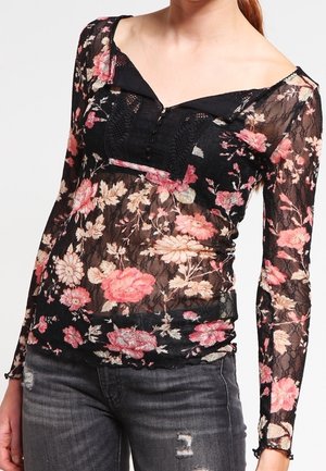 Femme portant un haut noir transparent à manches longues avec imprimé floral rose et beige et un jean gris foncé déchiré.