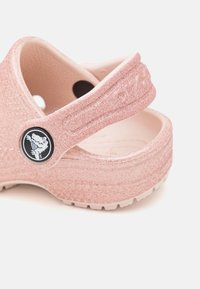 Różowe klapki z brokatem, z paskiem z tyłu, o formowanym kształcie i okrąłym logo. Teksturowana powierzchnia i lekki materiał.