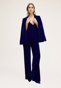 Completo in velluto a due pezzi di un blu profondo. Presenta un top aderente con scollatura a V e pantaloni a gamba larga. Le maniche a mantella aggiungono dettaglio e dimensione.