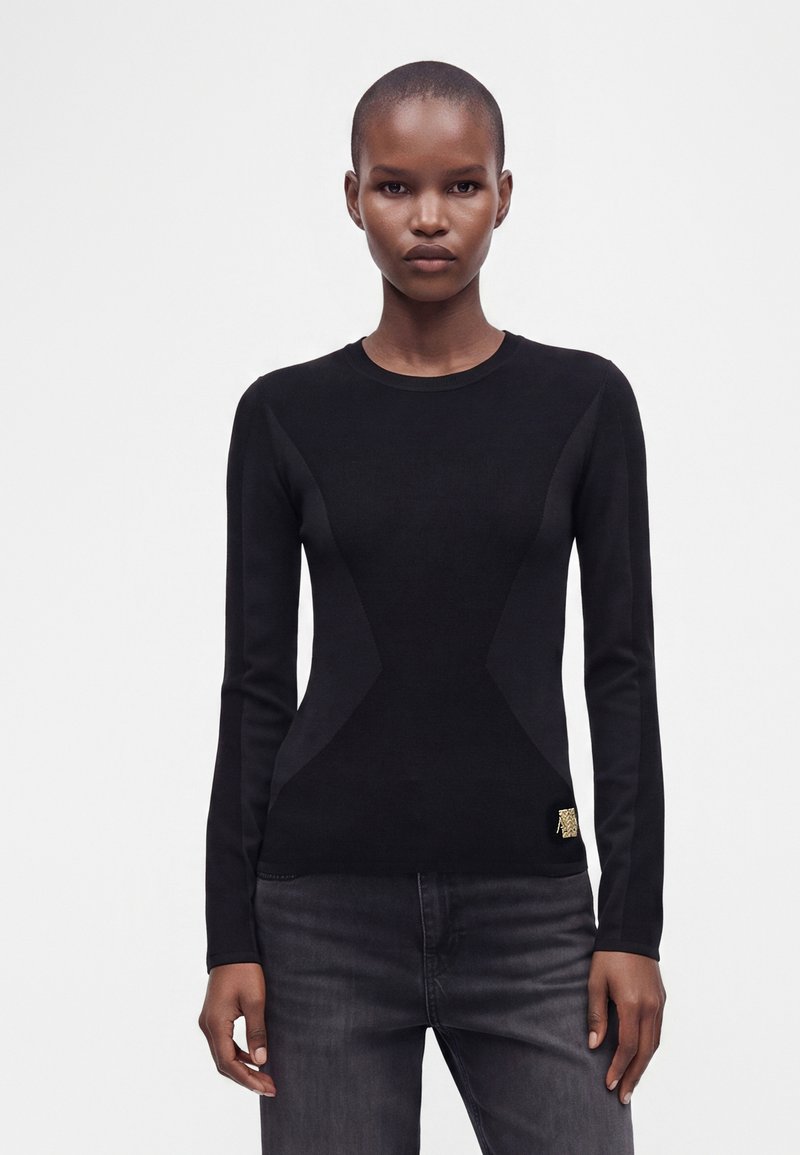 Personne aux cheveux courts portant un haut noir ajusté à manches longues et un jean gris foncé, debout devant un fond clair uni.