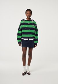 Groene en marineblauwe gestreepte trui met versieringen, gedragen over een witte blouse met kraag, gecombineerd met een denim rok en donkere schoenen.
