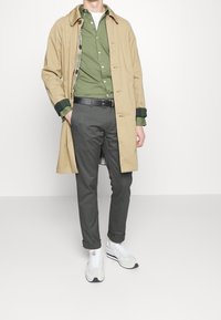 Beige trenchcoat över en grön skjorta med knappar, grå byxor och vita sneakers. Kappan har ett rutigt foder och uppvikta ärmar.