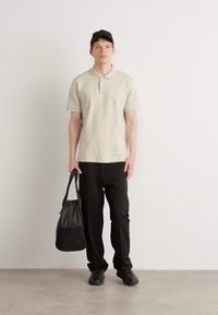 Svetlo siva polo majica s malim džepom, kombinovana sa crnim pantalonama i tamnosivom torbom na ramenu. Model nosi crnu kapu i crne cipele.