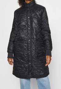 Cappotto lungo trapuntato nero con bottoni a pressione, collo alto e due tasche laterali; presenta un tessuto liscio e lucido e polsini a coste.