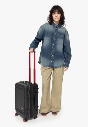 Black hard-shell suitcase with red telescopic handle, textured finish, and four wheels. Model wears a blue denim shirt and beige pants.