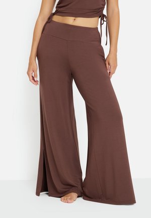 Donna che indossa pantaloni marroni a gamba larga con spacchi laterali e top corto abbinato, in piedi a piedi nudi su sfondo bianco.