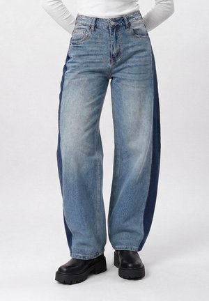 Personne portant un jean taille haute à jambes larges avec des panneaux latéraux foncés et des bottes noires épaisses, debout devant un fond clair uni.