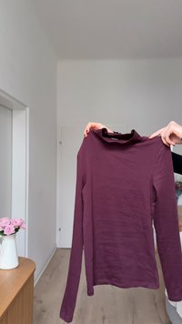 Dolga majica z dolgimi rokavi, oprijeta, v globoki vijolični barvi, rebraste teksture, z visokim ovratnikom. Postavljena na nevtralnem ozadju z minimalnimi vidnimi detajli.