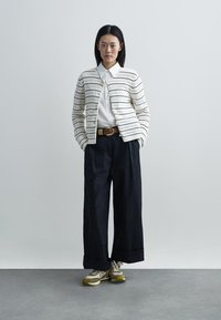 Cardigan lavorato a maglia con strisce orizzontali in crema e nero, camicia bianca, pantaloni neri larghi e sneakers beige con accenti oliva.