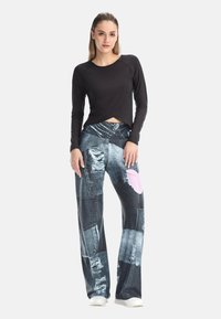 Kvinna klädd i en svart långärmad topp och vida byxor med ett patchworkmönster av riven denim, samt vita sneakers, stående mot en grå bakgrund.