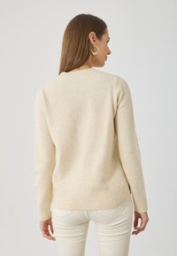 Suéter de color beige claro, con textura acanalada, mangas largas y escote redondeado. Textura suave, diseño minimalista, vista posterior con un ligero dobladillo caído.