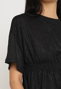 Sort kortærmet top med en samlet talje, fremstillet af et skinnende, tekstureret stof, der reflekterer lys og skaber en subtil glimmer-effekt.