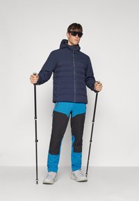 Μπλε επενδυμένο μπουφάν με κουκούλα, μαύρο και μπλε παντελόνι πεζοπορίας, αθλητικές μπαστούνες και ανοιχτό γκρι παπούτσια, σε ουδέτερο φόντο.