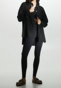 Camicia nera con texture e bottoni abbinata a leggings neri. Sandali marroni con dettaglio a fibbia. Messa a fuoco sull'aspetto stratificato dell'outfit.