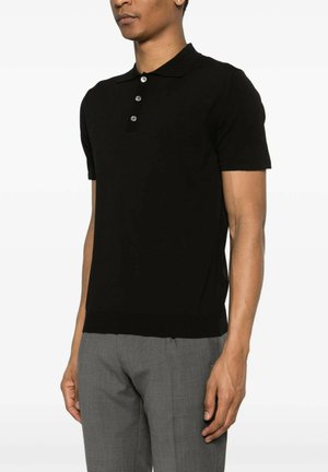 Uomo che indossa una polo nera a maniche corte con tre bottoni e pantaloni grigi su misura, in piedi contro uno sfondo bianco.