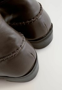 Stivali alla caviglia in pelle marrone con un design cucito lungo le cuciture e un tacco nero basso. Texture liscia con una forma arrotondata.