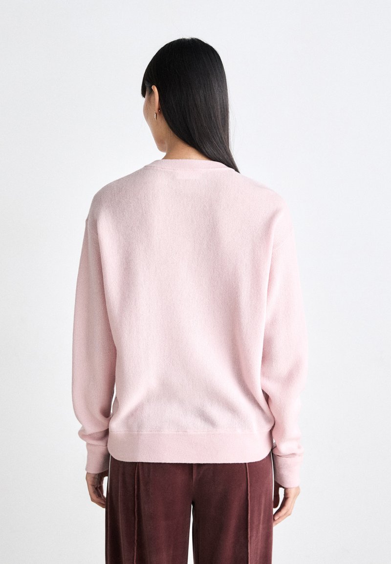 Pull rose en tissu doux et texturé, avec une coupe décontractée, des poignets côtelés et des détails de couture au dos. Porté avec un pantalon rouge foncé.