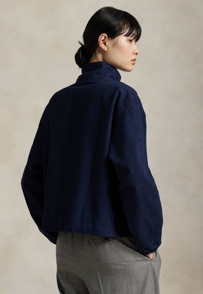 Kvinde med mørkt hår sat tilbage iført en navyblå jakke og grå bukser, stående i profil med hænderne i lommerne mod en neutral baggrund.