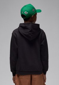 Must kapuutsiga pusa, millel on kenguruvets ja nööriga kapuuts, kombineeritud rohelise nokamütsiga, millel on tikitud logod, nähtud tagantpoolt.