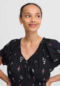 Robe noire avec un décolleté en V et des manches courtes bouffantes, comportant un motif floral en blanc et rose. Corsage smocké texturé et tissu doux.