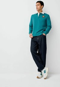 Tyrkysová polokošile s dlouhým rukávem a kontrastním bílým límečkem, s oranžovým vyšitým logem, v kombinaci s tmavými denimovými kalhotami a bílými teniskami.