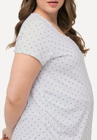 Camiseta blanca de manga corta, con lunares azules. La tela parece suave, con un detalle de fruncido lateral para un ajuste ajustable.