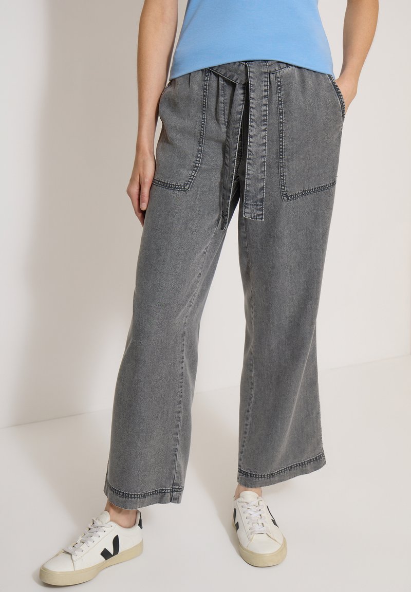 Personne portant un pantalon large gris délavé avec de grandes poches avant et une ceinture assortie, associé à des baskets blanches et un haut bleu clair.