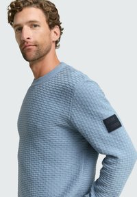 lichtblauwe gestructureerde trui met een ronde hals, lange mouwen en een zwart stoffen label op de linkerschouder.