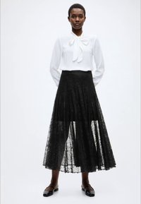 Λευκό πουκάμισο με φτιαγμένη φιόγκο, σε συνδυασμό με μαύρη δαντελένια πλισέ φούστα που φτάνει ως τους αστραγάλους και μαύρα παπούτσια με επίπεδη σόλα.