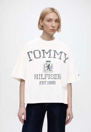 Young person with short blonde hair wears oversized white Tommy Hilfiger T-shirt with large logo and navy pants against plain background.
