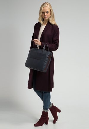 Borsa a tracolla in pelle nera con doppi manici, forma rettangolare e chiusura a zip. Indossata da una persona con un cappotto bordeaux e jeans blu.