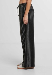 Černé pruhované kalhoty s širokými nohavicemi a stahovacími pásky v pase, s kovovými detaily a jemnou texturou. Nosené s lehkou obuví.