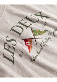 Grå bomullssweatshirt med grønn og rød brodering. Designet inkluderer flagg og tekst som leser "LES DEUX" og "International Sporting Goods."