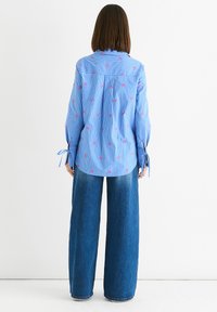 Frau mit schulterlangem braunem Haar, die ein blaues gestreiftes Shirt mit pinken Vögeln und weiten blauen Jeans trägt, vor einem weißen Hintergrund stehend.
