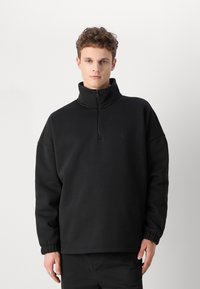 Felpa nera a collo alto con chiusura a mezza zip. Realizzata in un tessuto liscio, presenta una vestibilità ampia e polsini elasticizzati.