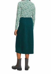 Femme portant un haut à manches longues vert à motifs, une jupe midi taille haute vert foncé et des bottines noires, vue de dos.