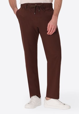 Uomo che indossa pantaloni marrone scuro con coulisse e sneakers bianche, in piedi con una mano in tasca e una mano rilassata.