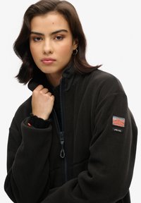 Giacca in pile nera con colletto alto, chiusura con zip e patch con logo sulla manica. Presenta un tessuto testurizzato e un interno a contrasto.