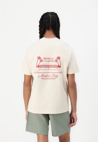 T-shirt beige a maniche corte con un design grafico rosso di palme e un campo da tennis, con il testo "Monte Carlo Country Club" e "Solo per membri."