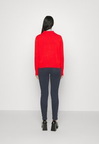 Czerwony sweter z wysokim kołnierzem, z prążkowanymi mankietami i dołem. Połączony z dopasowanymi ciemnoniebieskimi dżinsami i czarnymi butami na dużym obcasie.