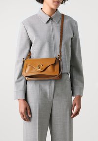 Brown leather handbag with a curved flap and gold hardware clasp. Compact design with a shoulder strap and contrasting accents. Check-patterned outfit.