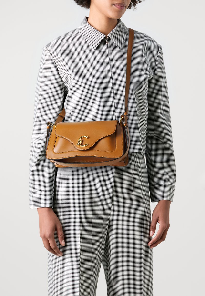 Brown leather handbag with a curved flap and gold hardware clasp. Compact design with a shoulder strap and contrasting accents. Check-patterned outfit.