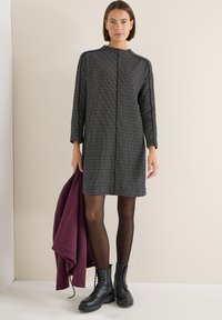 Černé a bílé šaty s vzorem, dlouhými rukávy, vysokým výstřihem a rovný střih, doplněné černými bojovými botami a fialovou bundou.