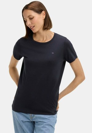 Femme regardant vers le bas, portant un t-shirt noir uni à manches courtes et un jean bleu clair, debout les mains derrière le dos.