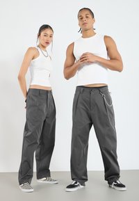 Graue weit geschnittene Hose mit glatter Textur und Kettendetail. Kombiniert mit weißen, eng anliegenden Tanktops und Sneakers.