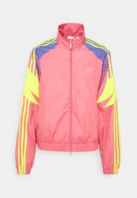 Ljusröd sportjacka med dragkedja, gula ränder p�å ärmarna, lila axelaccents och en liten gul logotyp på bröstet.