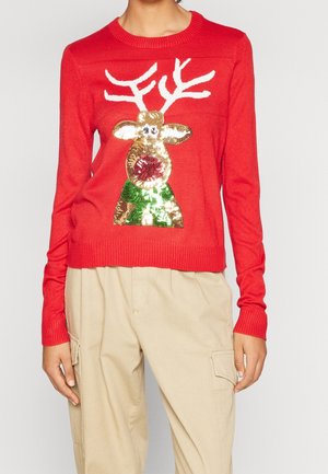 Persona che indossa un maglione rosso con una renna di paillettes dorate e verdi e pantaloni beige con tasche laterali.